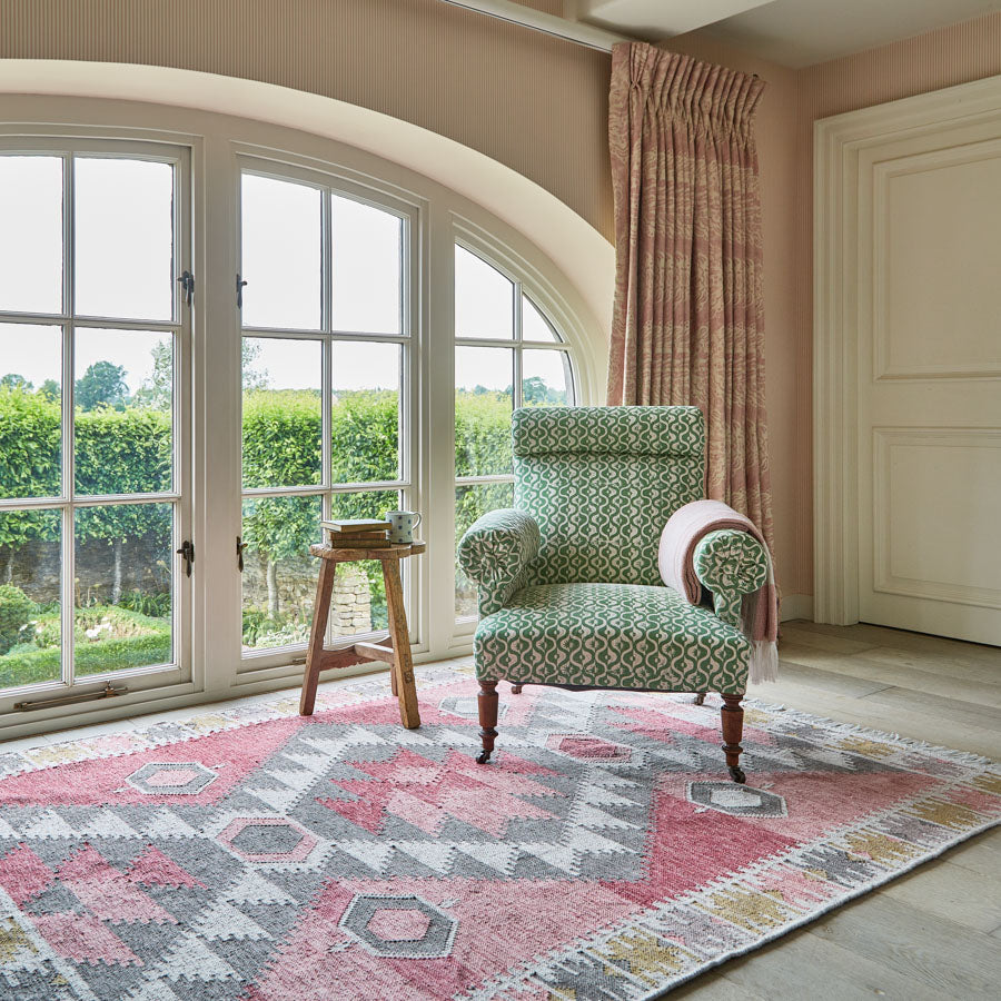 A geometric patterned rug in multiple colors placed on a floor in a room with a chair, with a view of greenery outside through the window.