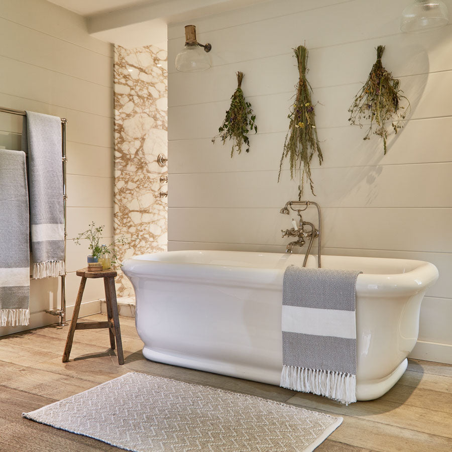 A soft grey chenille rug with white zig-zag stripes, placed on a wooden floor next to a white bathtub and under a wall-mounted lamp.