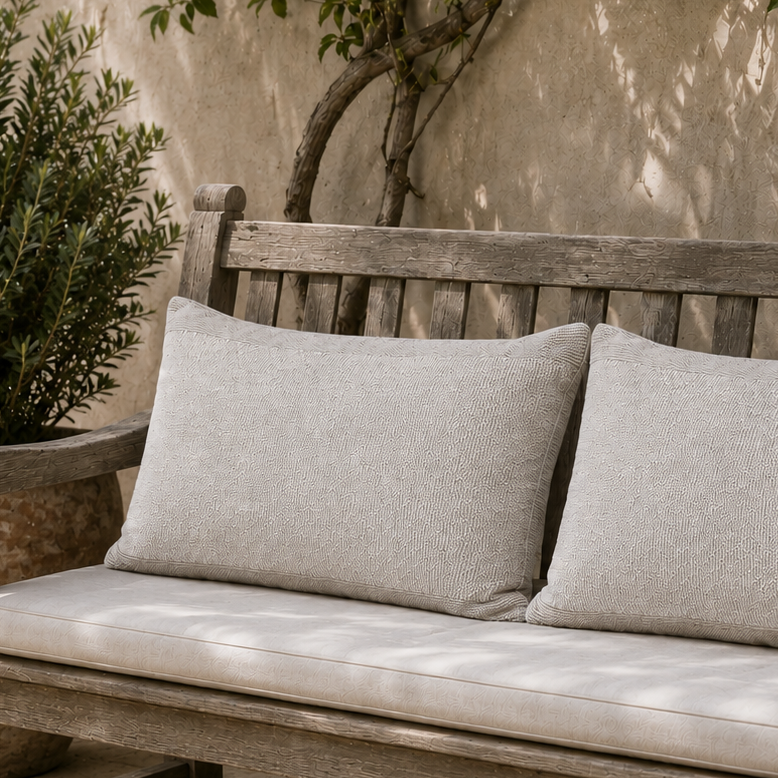Wooden bench with gray cushions in an outdoor setting with plants and stone wall.