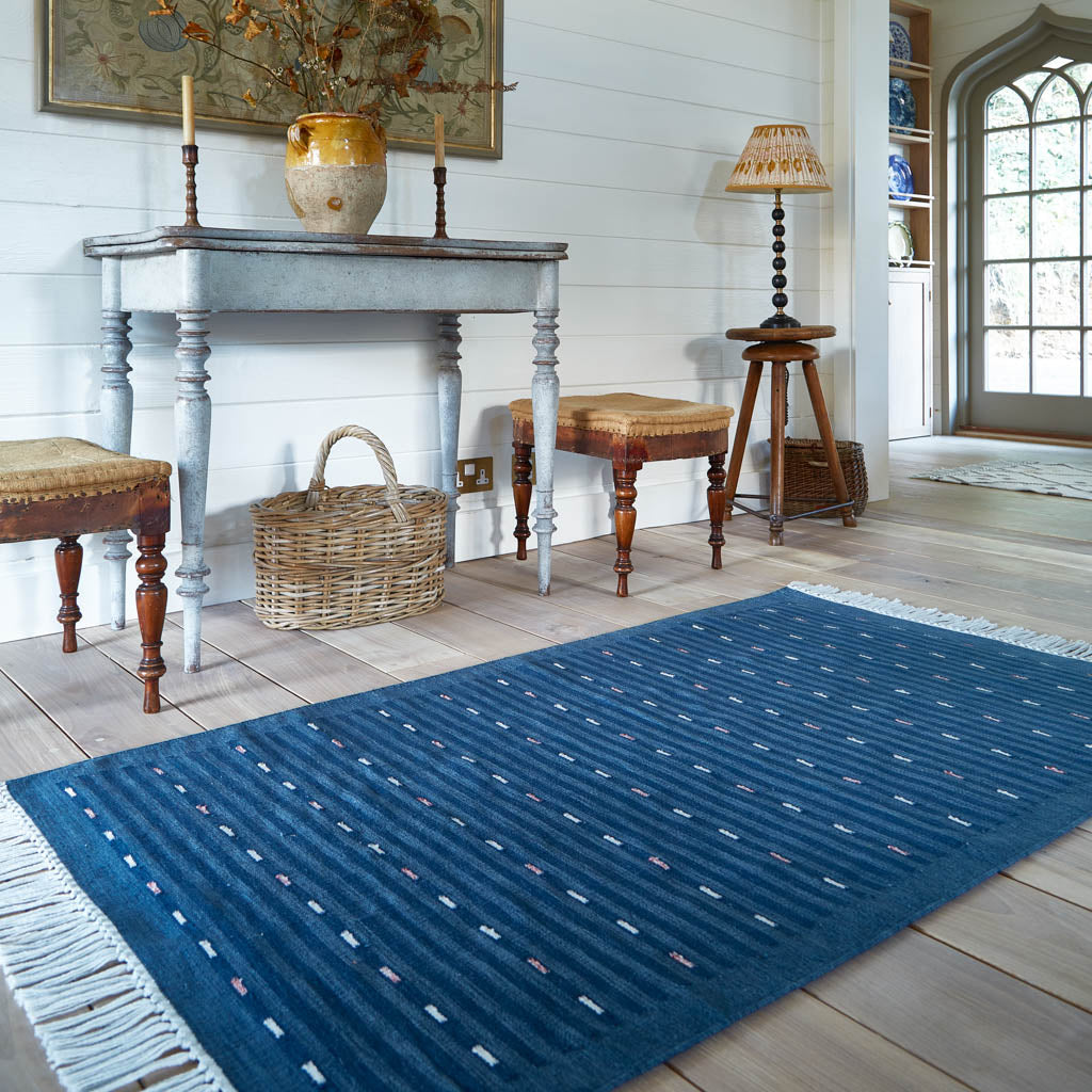 A Cosmo Indigo Rug with a bohemian striped design in faded indigo blue, placed on a floor in a room with furniture around it.