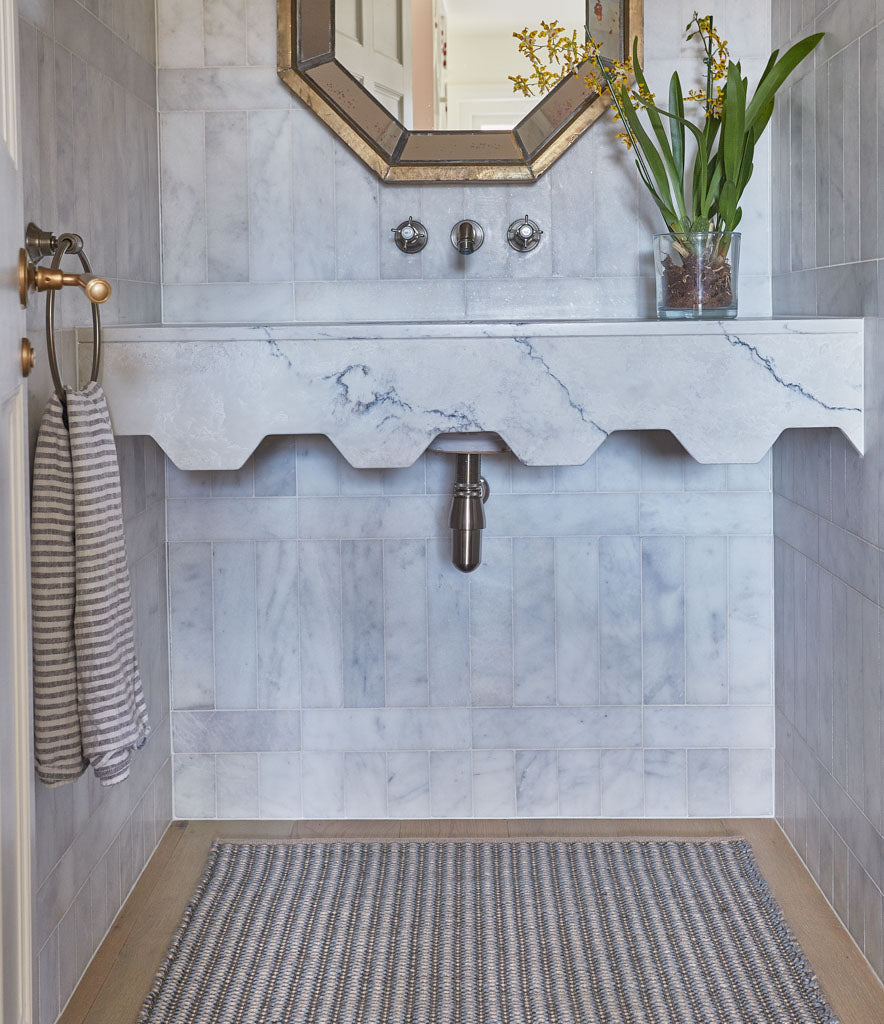 A striped rug with sky blue and charcoal colors, placed on a bathroom floor with a white marble wall and a wooden cabinet.