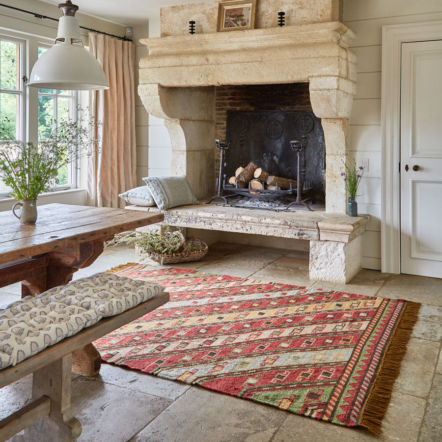 A multicolored geometric patterned rug with a tassel, placed on a stone floor in front of a fireplace in a room with wooden beams and a bench.