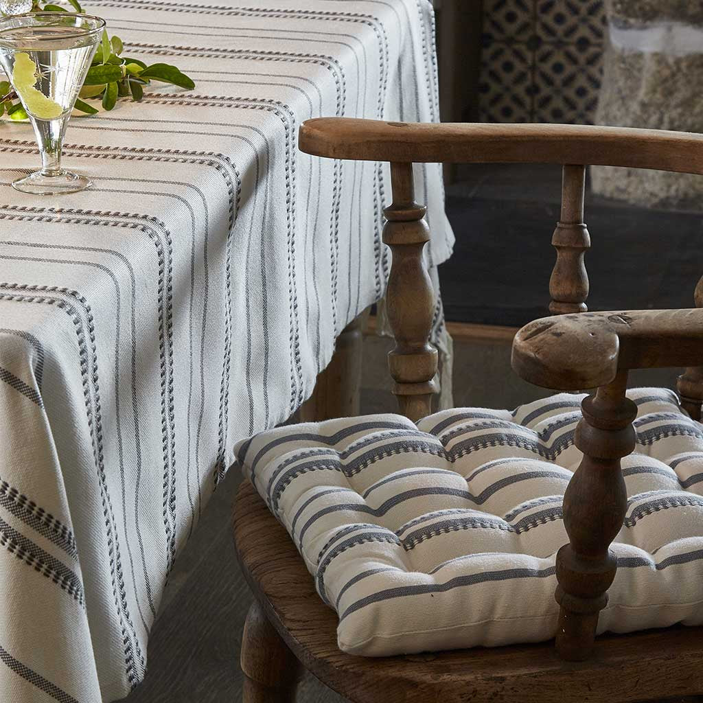 A blue chair cushion with a striped pattern placed on a wooden chair, with a glass of white wine and a striped tablecloth in the background.