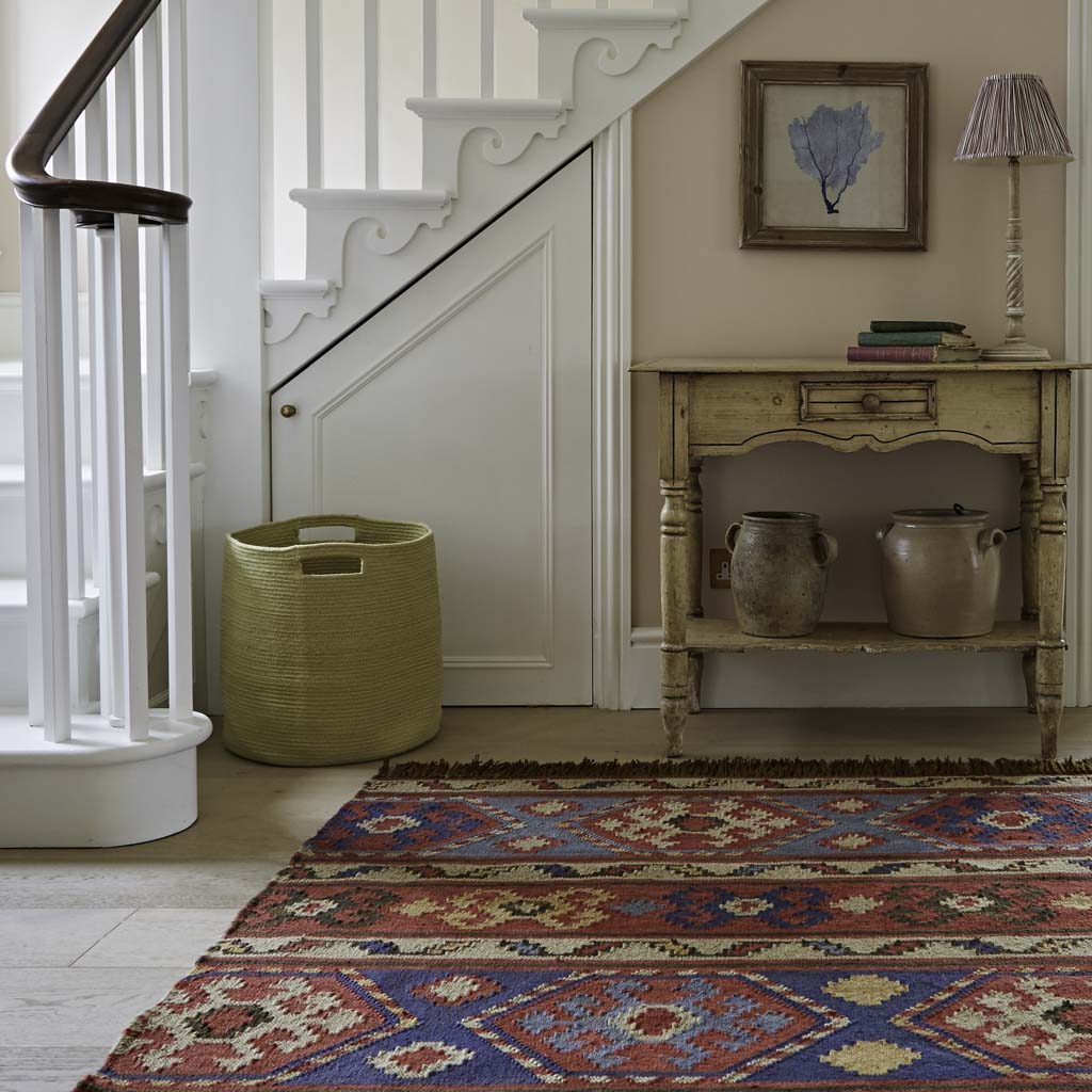 A multicolored geometric patterned rug placed on a floor, extending into a hallway, with a wooden side table and lamp, and a stairway in the background.