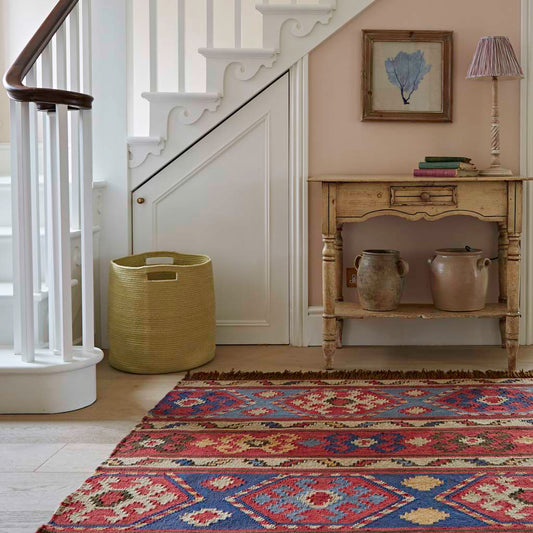 A multicolored geometric patterned rug placed on a floor, extending into a hallway, with a wooden side table and lamp, and a stairway in the background.