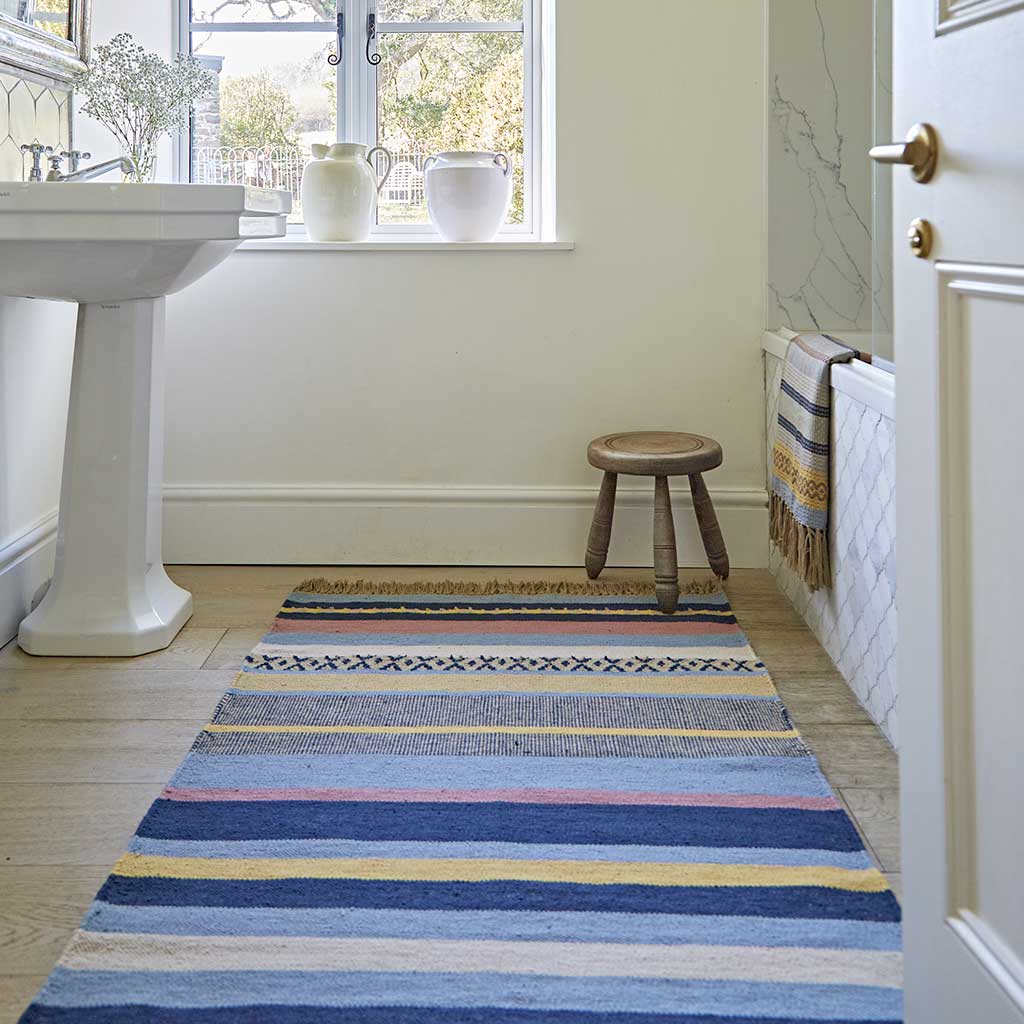 A multicolored striped rug with blue, pink, and gold hues laid out on the floor in a room with a white sink and wooden stool.